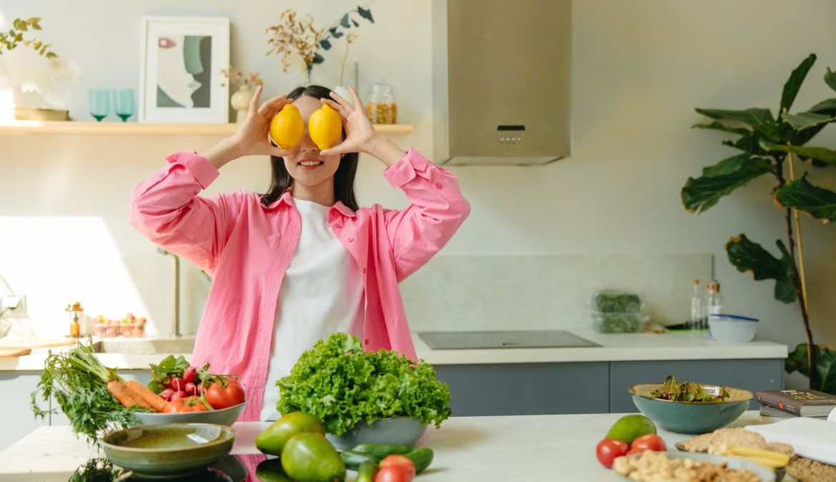 A woman playfully holding lemons over her eyes in a bright, lively kitchen setting filled with fresh vegetables.